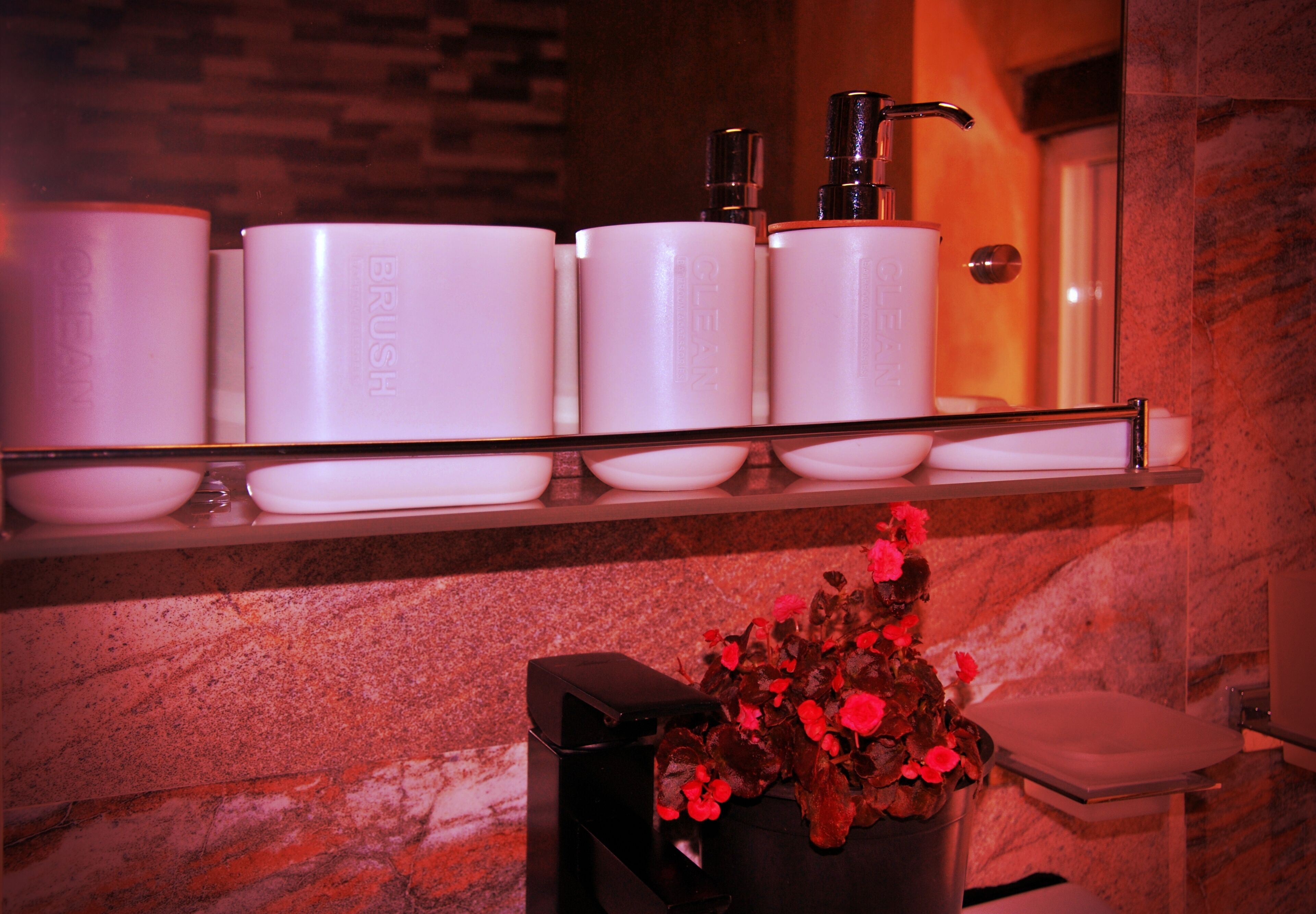 A bathroom with a marble wall, a shelf with toiletry containers, and a vase with flowers.