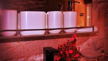 A bathroom with a marble wall, a shelf with toiletry containers, and a vase with flowers.