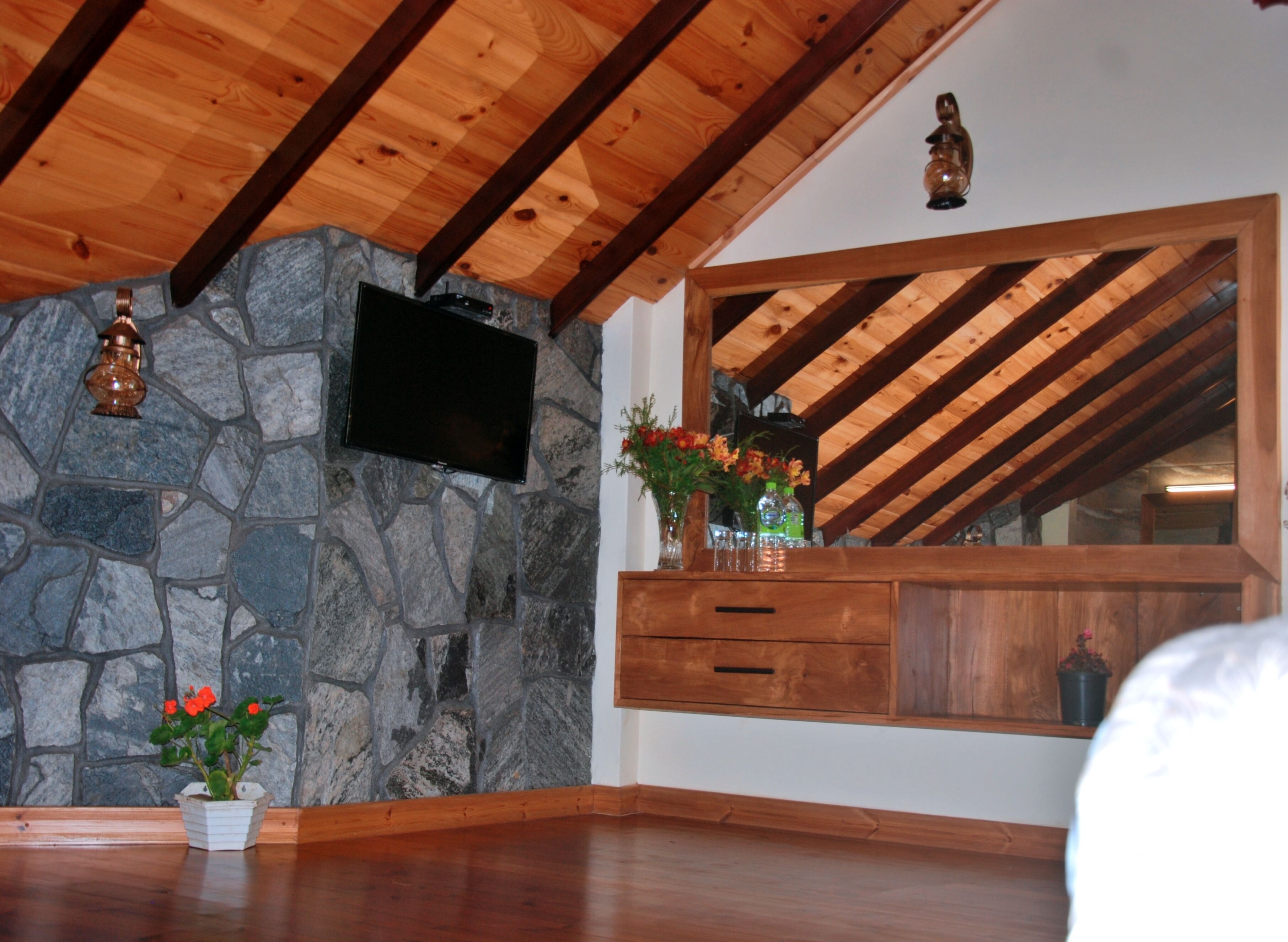 A room with a stone wall, wooden ceiling beams, a flat-screen TV mounted on the wall, a wooden cabinet with a mirror, and a potted plant.