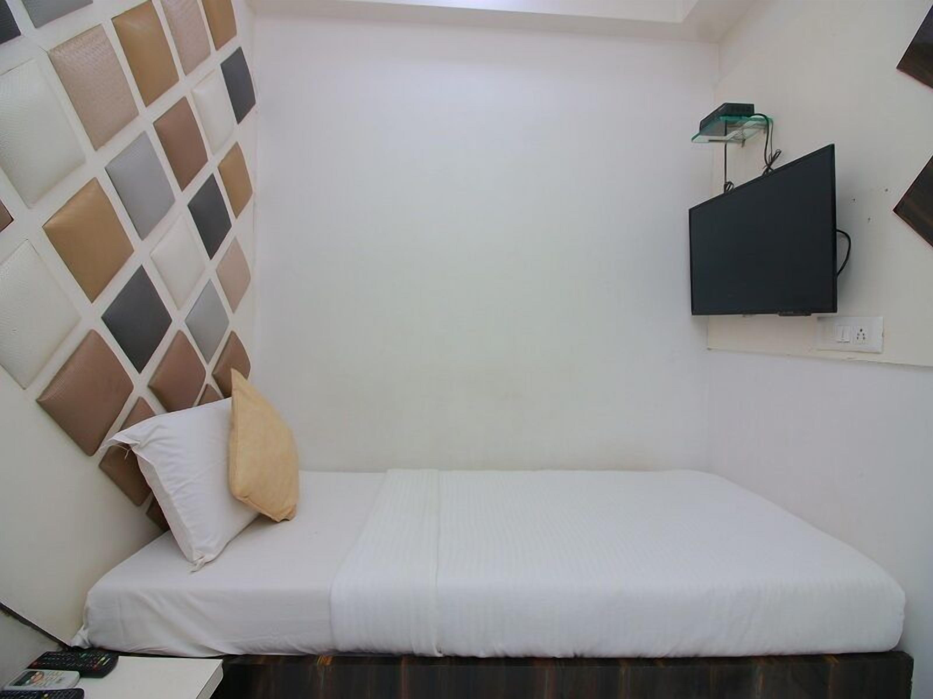 A small, single-bed room with a plain white wall, a mounted TV, and a simple bed with white bedding.