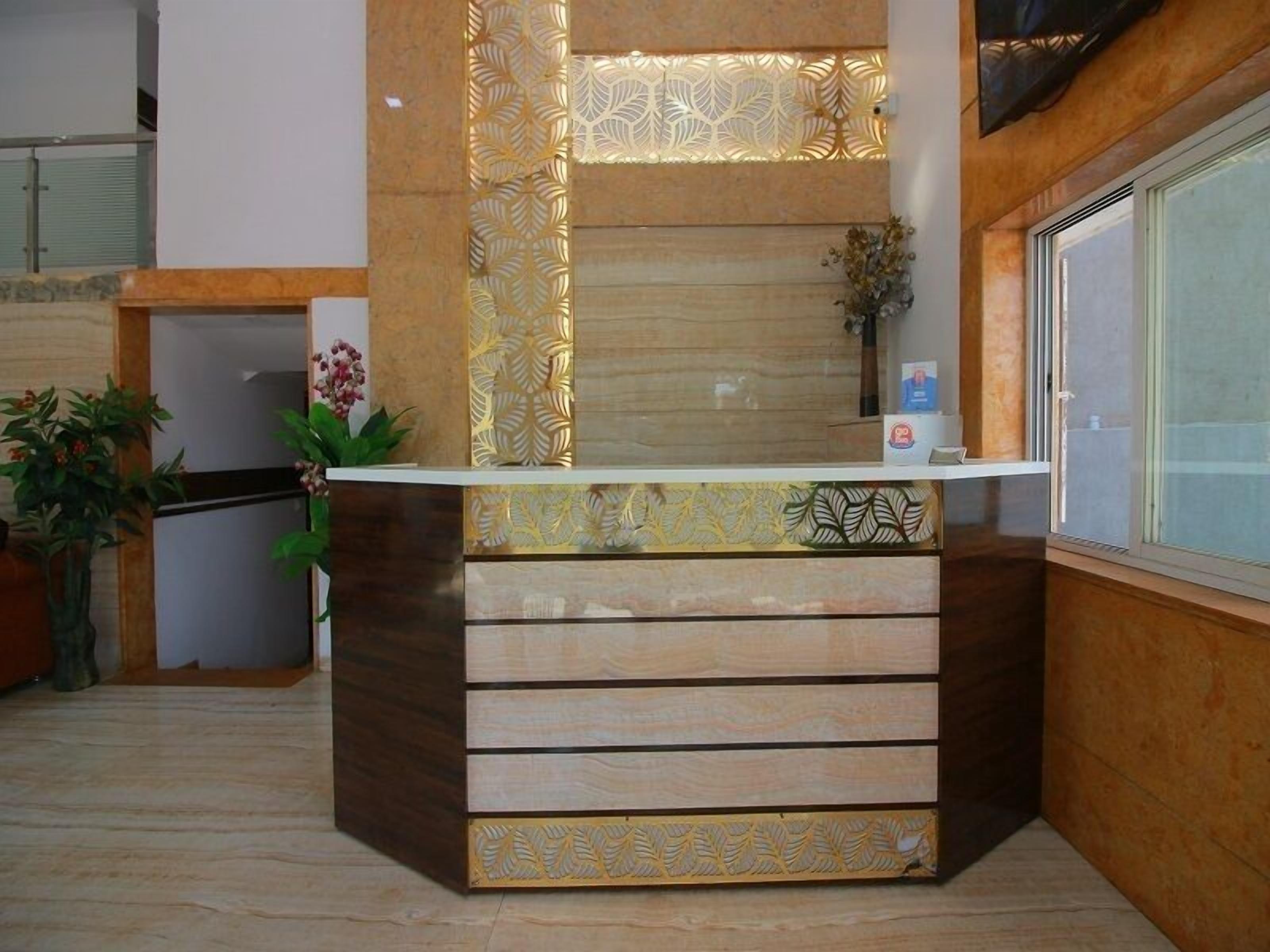Un mostrador de recepción con detalles de madera y motivos vegetales, flanqueado por una ventana y una planta.