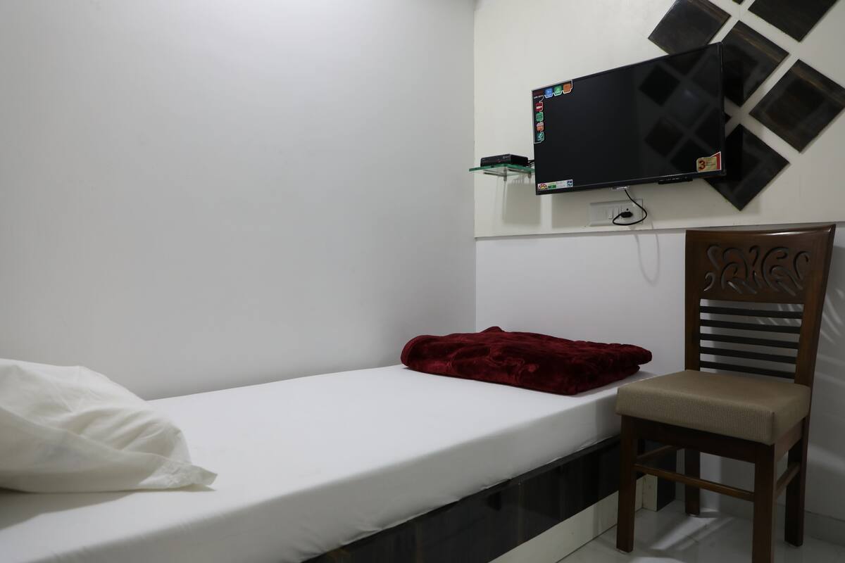 A small, single-bed room with a television mounted on the wall, a wooden chair with a cushion, and a red blanket on the bed.
