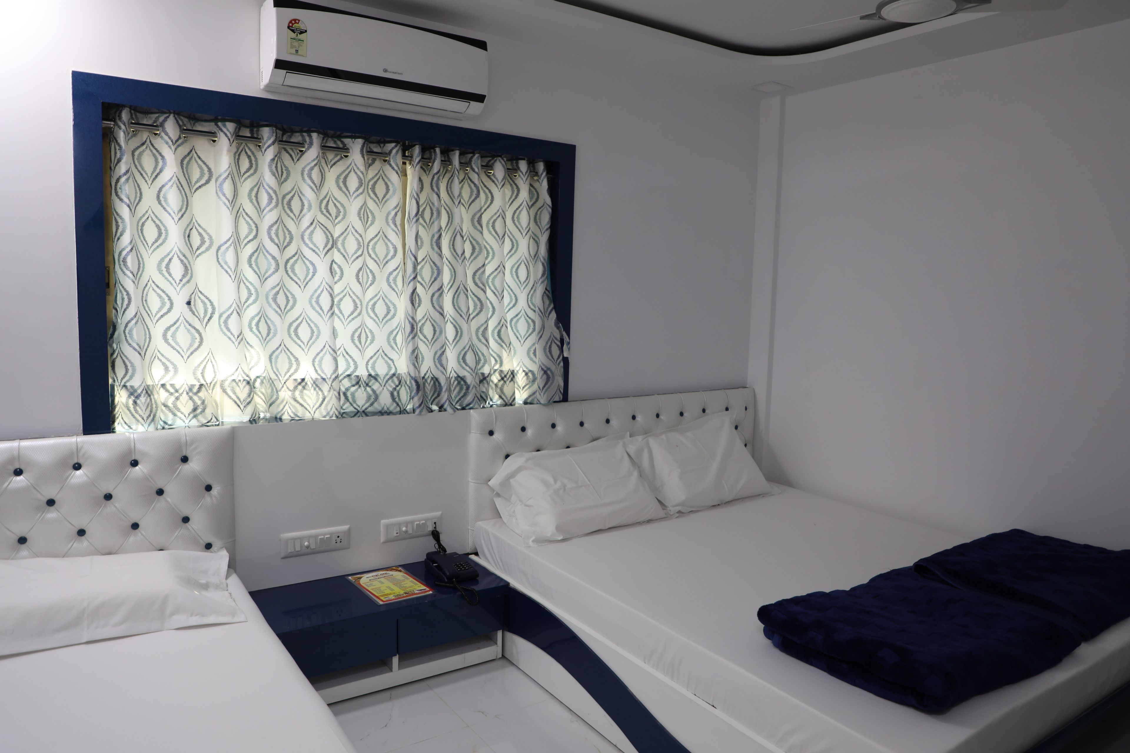 A bedroom with a bed, a bedside table, a power outlet, and a window with patterned curtains.