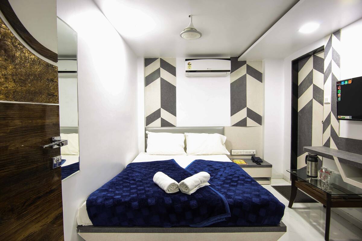 A modern hotel room with a neatly made bed, a bedside table with a lamp, and a flat-screen TV on a stand.