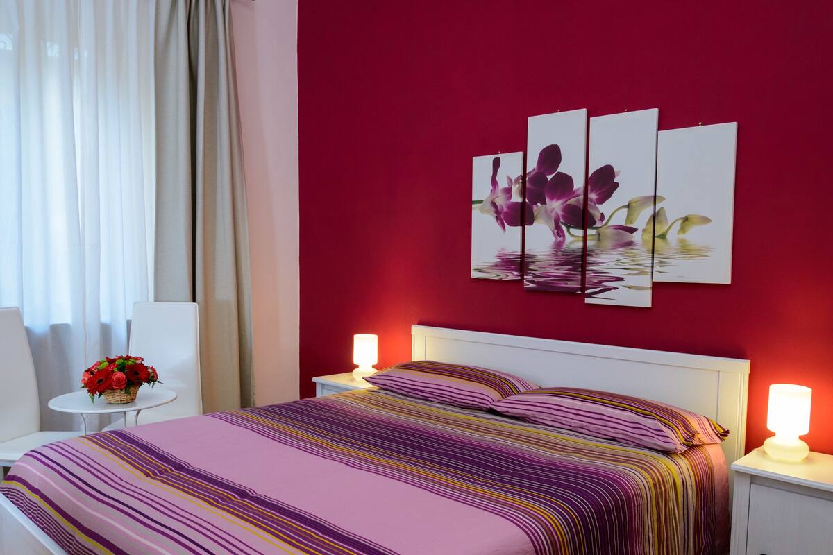 A bedroom with a bed, two bedside tables, a nightstand, a lamp, a chair, and a window with curtains.
