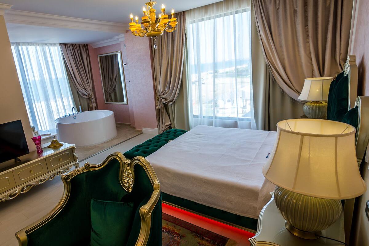A hotel room with a large bed, a sitting area with a green armchair, a chandelier, a bathtub, and a television.