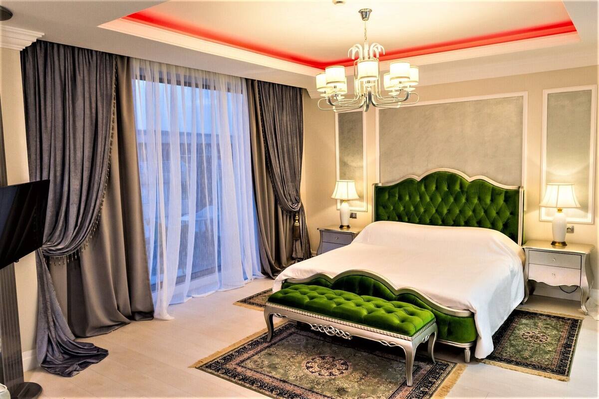 A hotel room with a large bed, a green upholstered bench, a chandelier, and a large window with curtains.