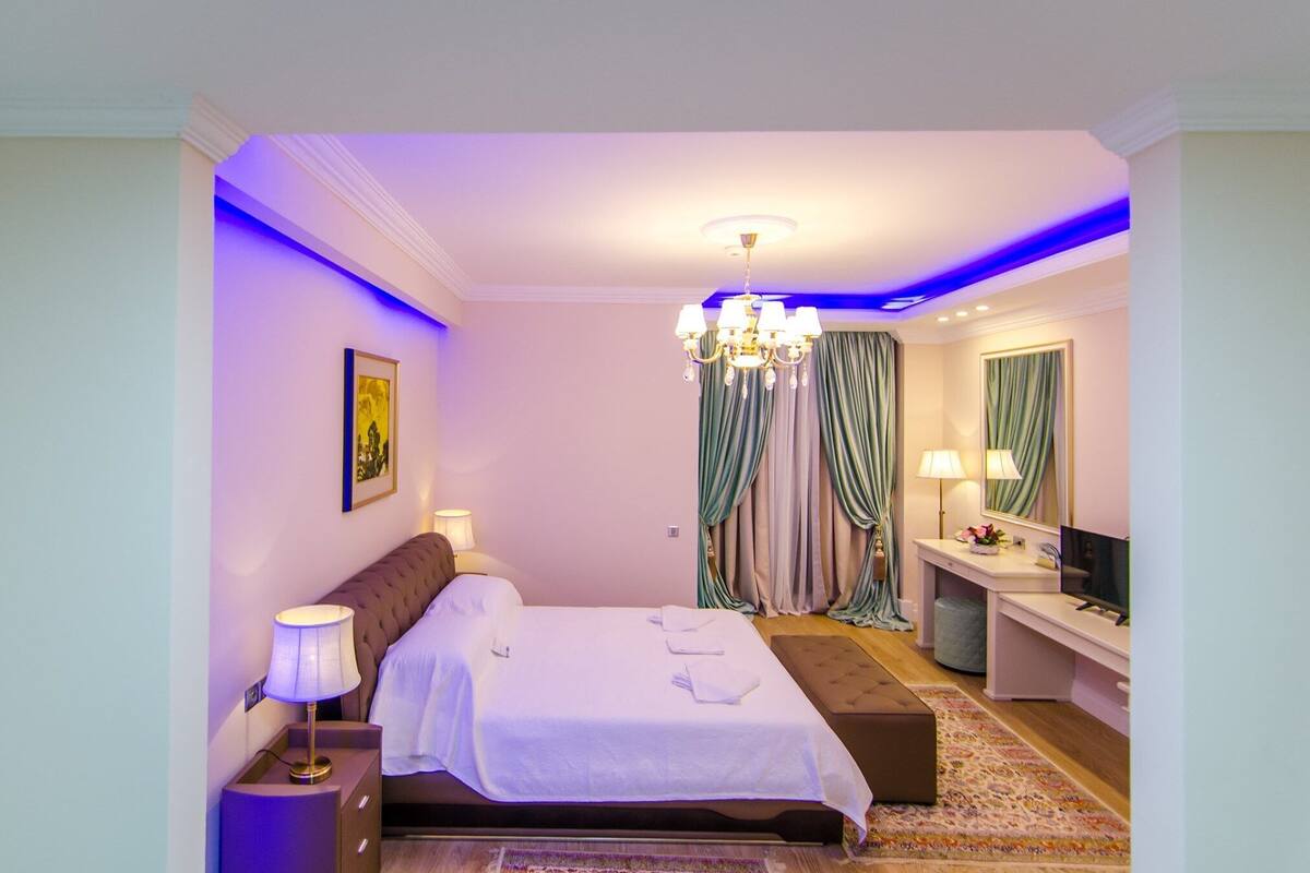 A hotel room with a bed, bedside table, lamp, TV, and a desk with a mirror.