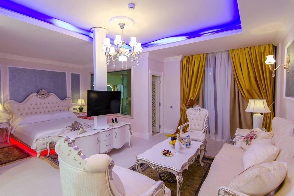 A luxurious bedroom with a large bed, a TV, a desk, and a dining area with a chandelier.