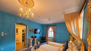 Habitación de hotel con dos camas, un candelabro, un televisor y vistas al exterior a través de una ventana con cortinas.