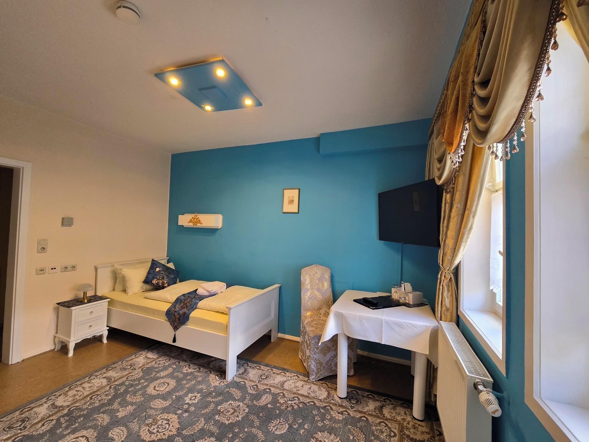 A bedroom with a bed, bedside tables, a desk, a chair, a television, and a patterned rug.