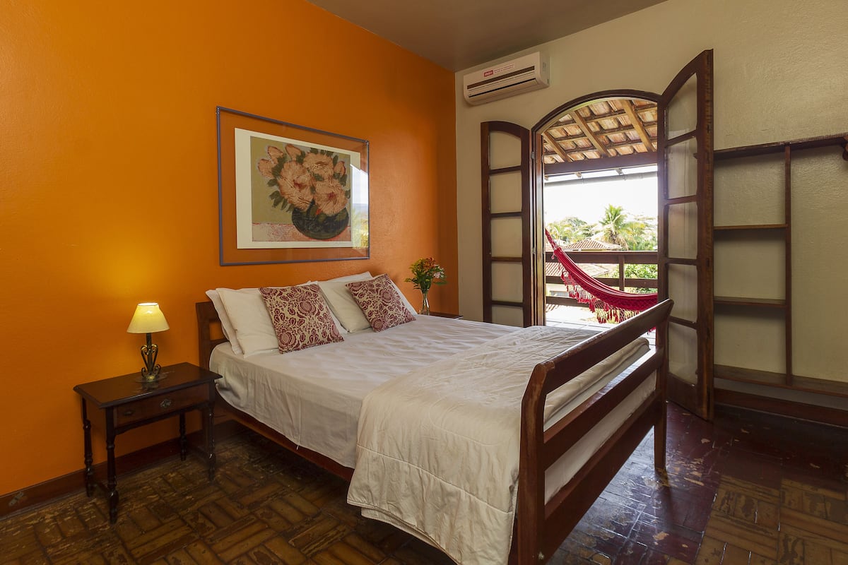 Une chambre à coucher comprenant un lit, une table de chevet, une lampe, un tableau encadré, un appareil de climatisation et une fenêtre donnant sur une chaise rouge et de la verdure à l’extérieur.