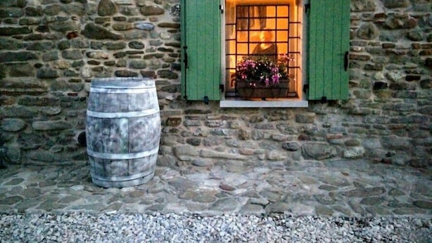 A stone wall with a window featuring green shutters, a metal barrel, and a flower box. - Azienda Agricola Baccagnano (Brisighella)