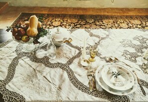Uma mesa posta com toalha de renda, uma tigela de frutas, um bule, uma vela e louças.