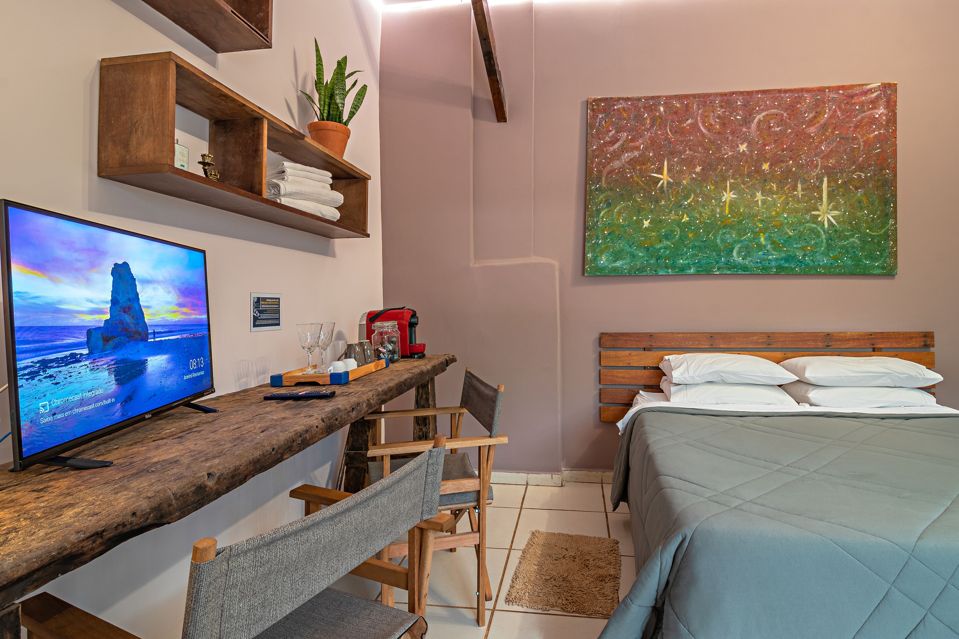 A bedroom with a bed, a desk with a computer monitor, a chair, a wooden shelf with plants, and a painting on the wall.