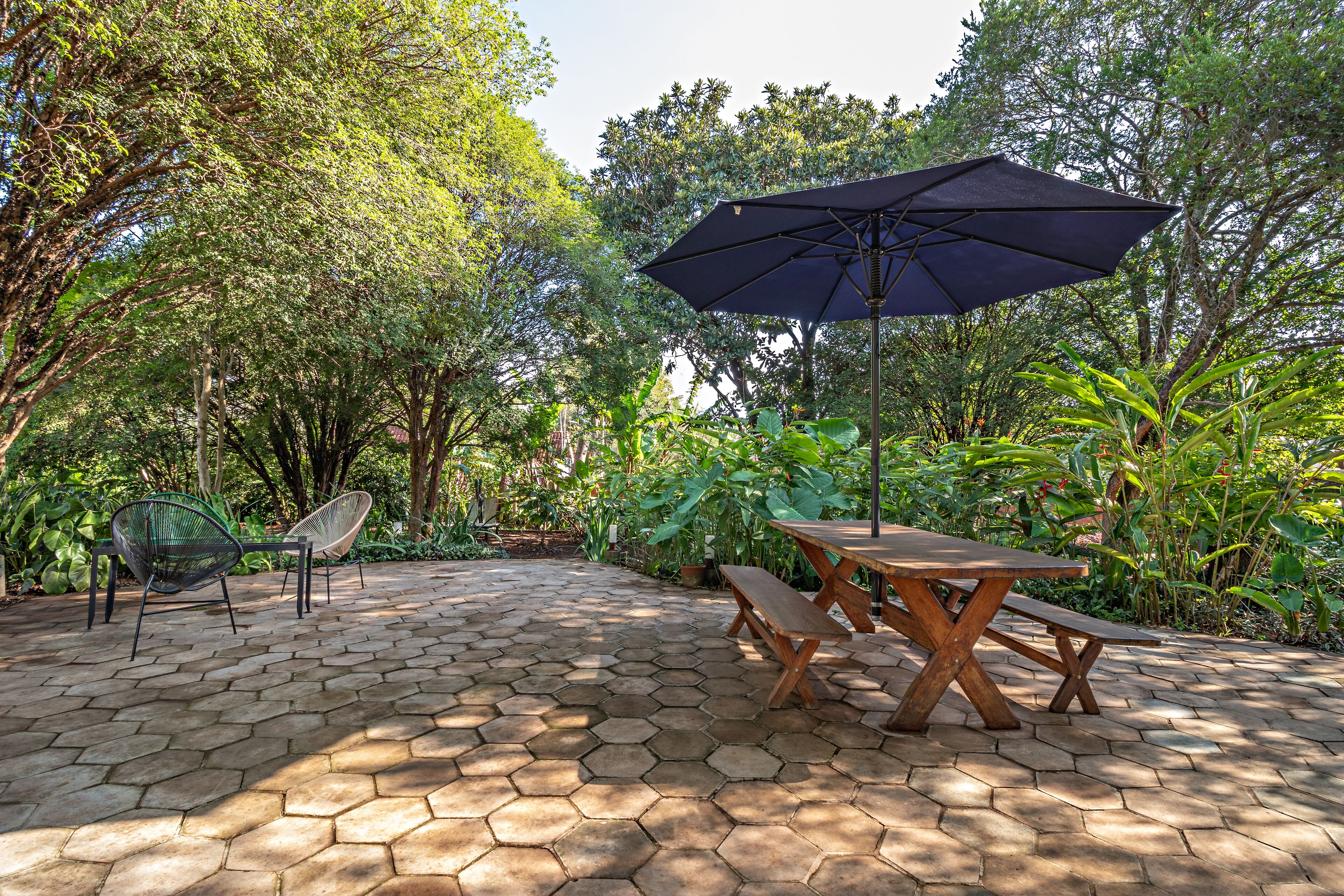 A hexagonal tiled patio with a wooden table and benches, surrounded by lush greenery and trees.