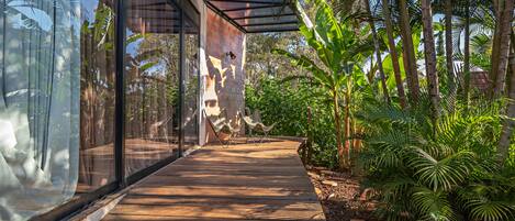 A wooden deck leading to a glass door, surrounded by tropical plants and a stone wall.