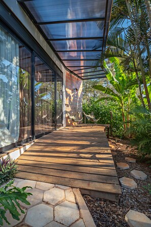 A covered outdoor area with a wooden deck, glass walls, and tropical plants. - Suítes no Quintal (Brotas)