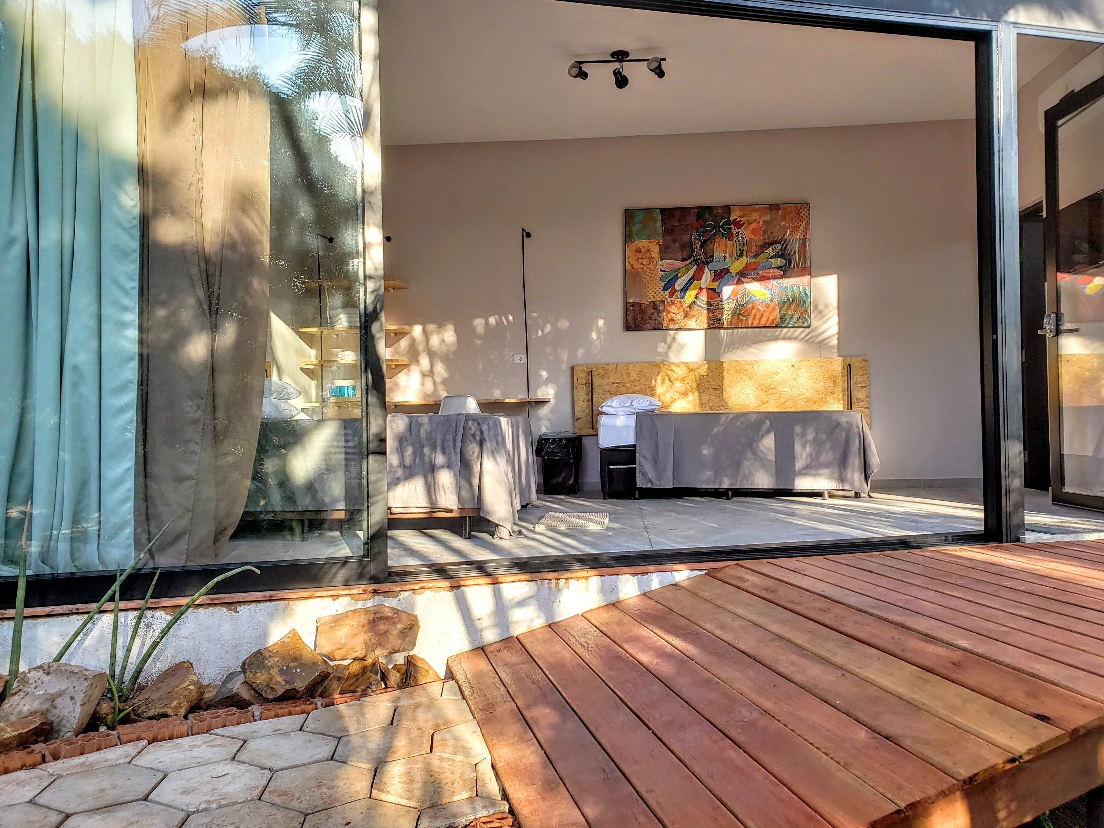 A modern room with a wooden deck, a large window with curtains, a painting on the wall, and a bed with a headboard.