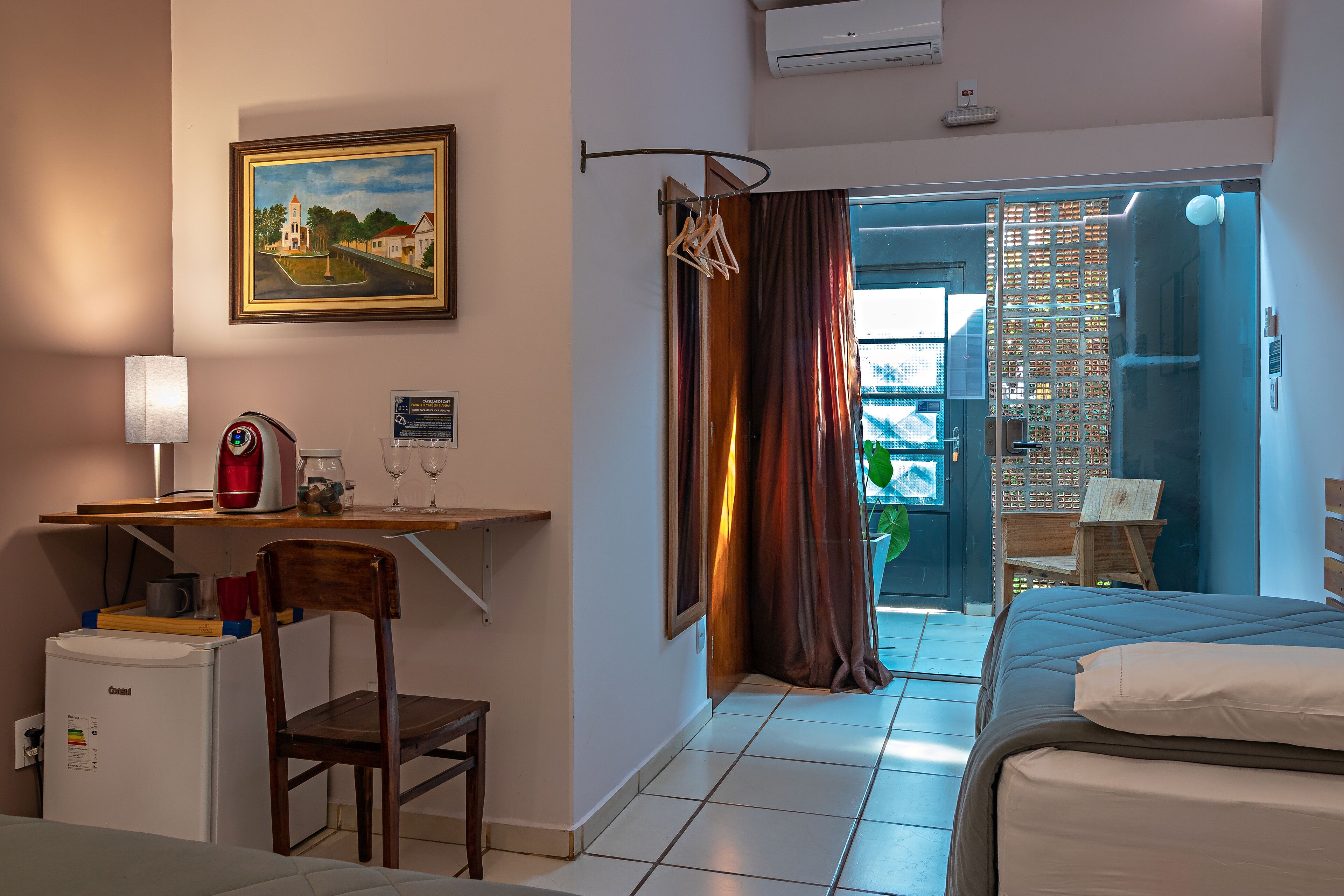 Une chambre d’hôtel avec un lit, un bureau doté d’une machine à café, une chaise, un réfrigérateur, un tableau accroché au mur et une vue à travers un rideau.
