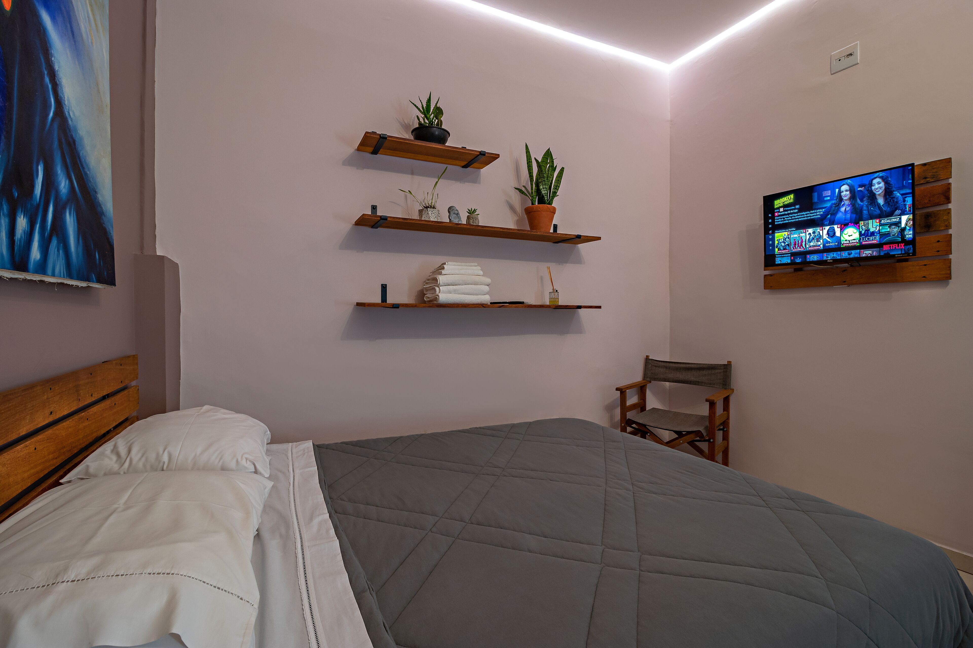 Een slaapkamer met een bed, een aan de muur bevestigde televisie en planken met planten en handdoeken.