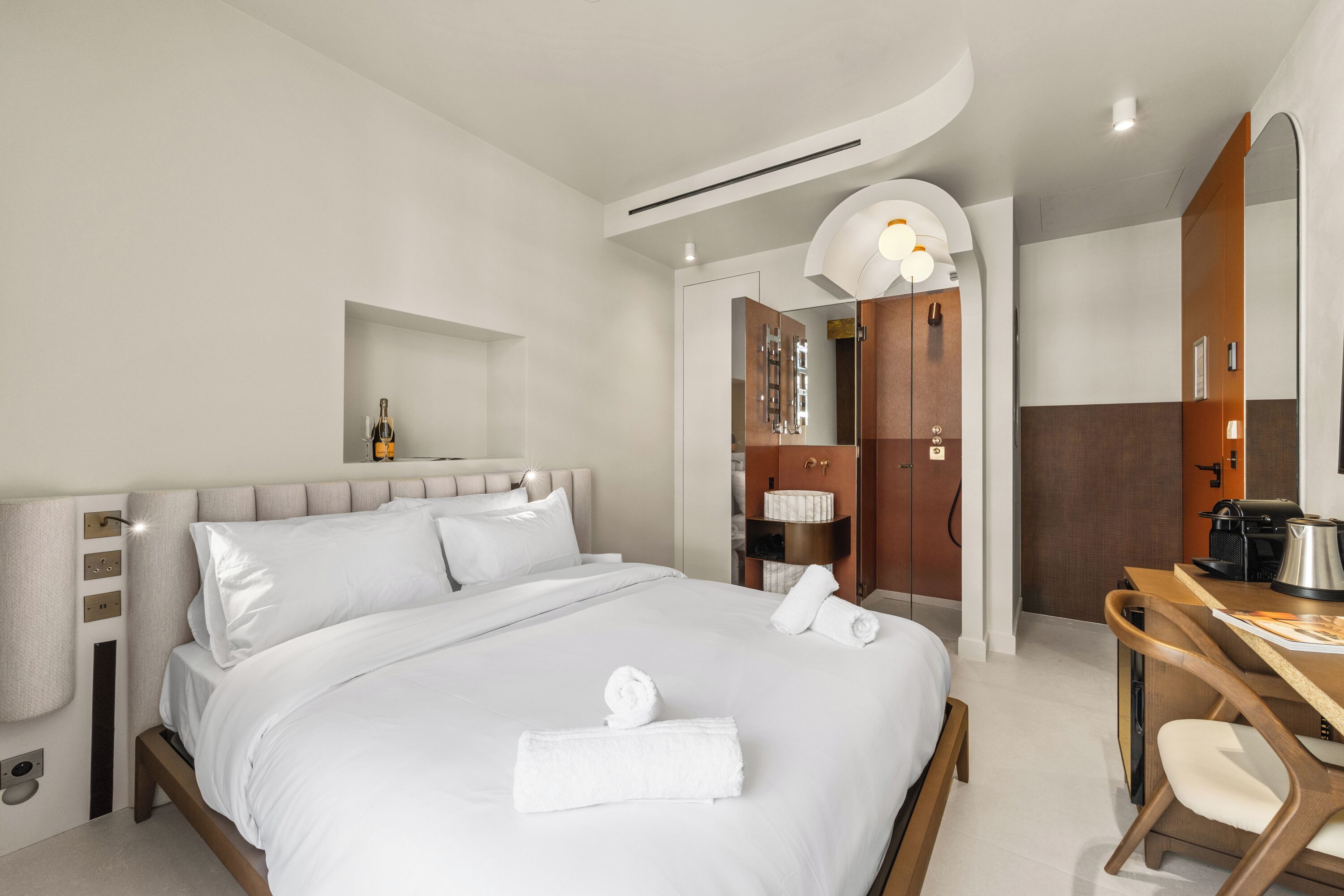 A modern hotel room with a large bed, a desk with a kettle, and a wardrobe.