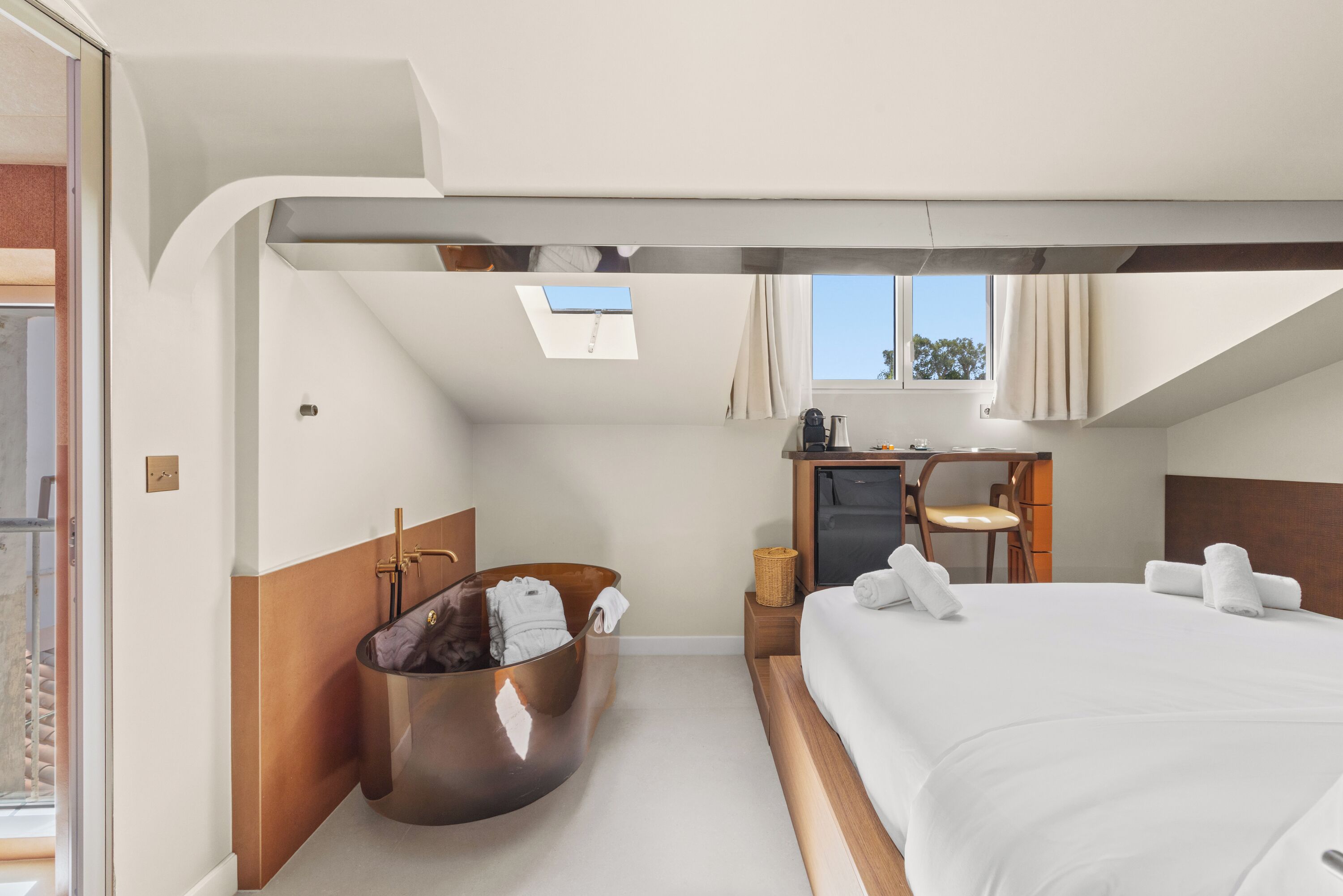 A modern hotel room with a large bed, a hanging bathtub, a desk, and a window.