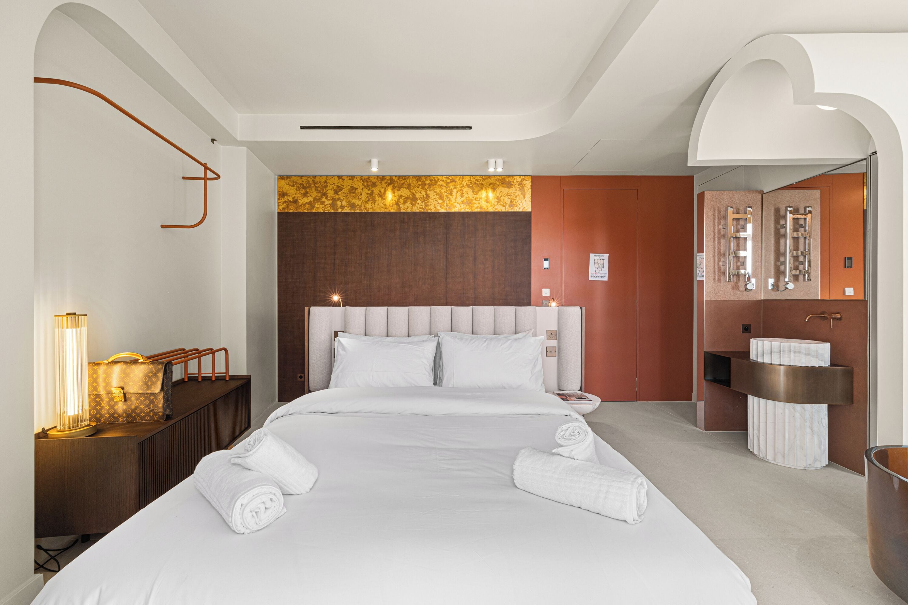 Ein modernes Hotelzimmer mit einem großen Bett, einem hölzernen Kopfteil, einem Nachttisch mit Lampe und einem Badezimmer mit Marmorwaschbecken.