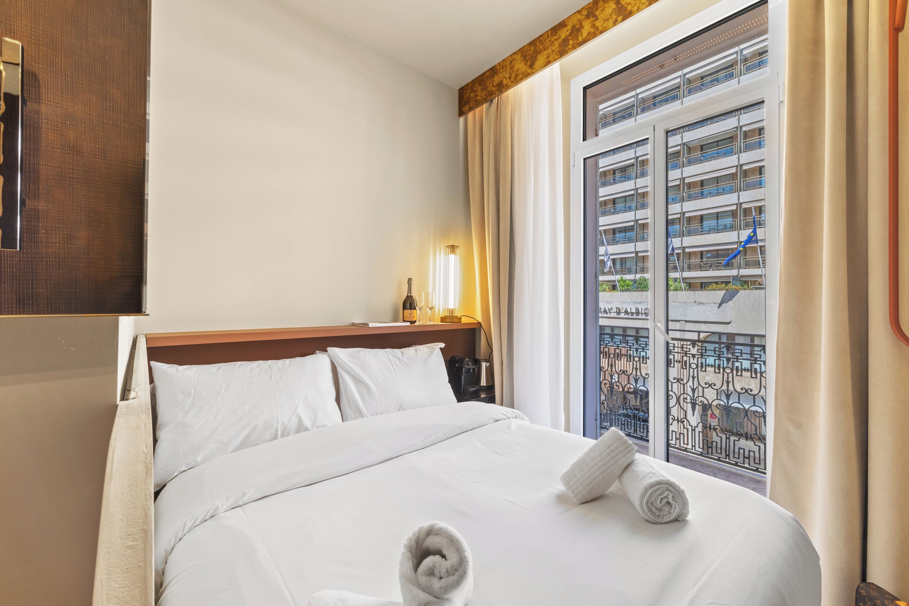 A hotel room with a bed, a nightstand, a lamp, and a window with a view of buildings.