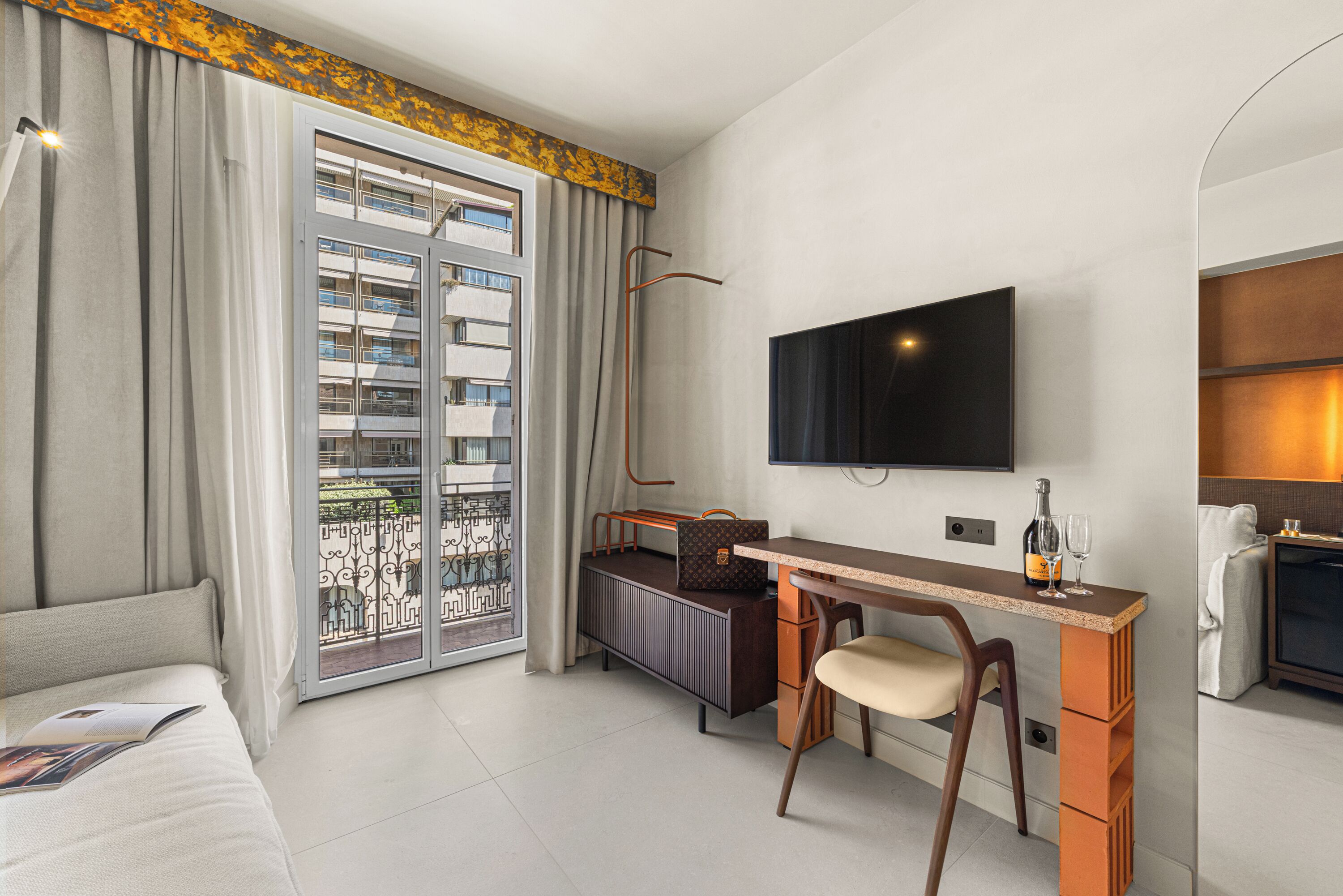Une chambre d’hôtel avec un lit, un bureau et une chaise, un téléviseur, un radiateur et une vue sur un immeuble à travers une porte vitrée.