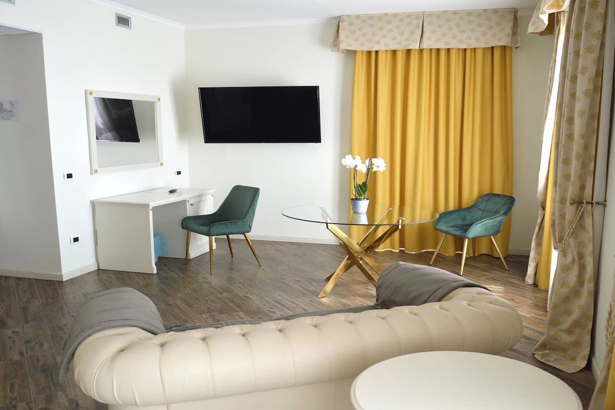 A hotel room with a sofa, a small round table, a desk with a chair, a TV mounted on the wall, and a window with curtains.