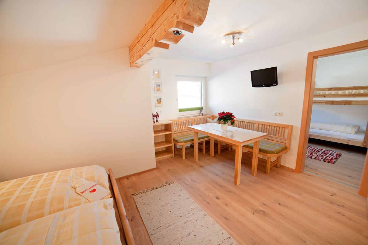 Um quarto aconchegante com uma cama, uma mesa de madeira com cadeiras, uma TV fixada na parede e uma janela que ilumina naturalmente o espaço.