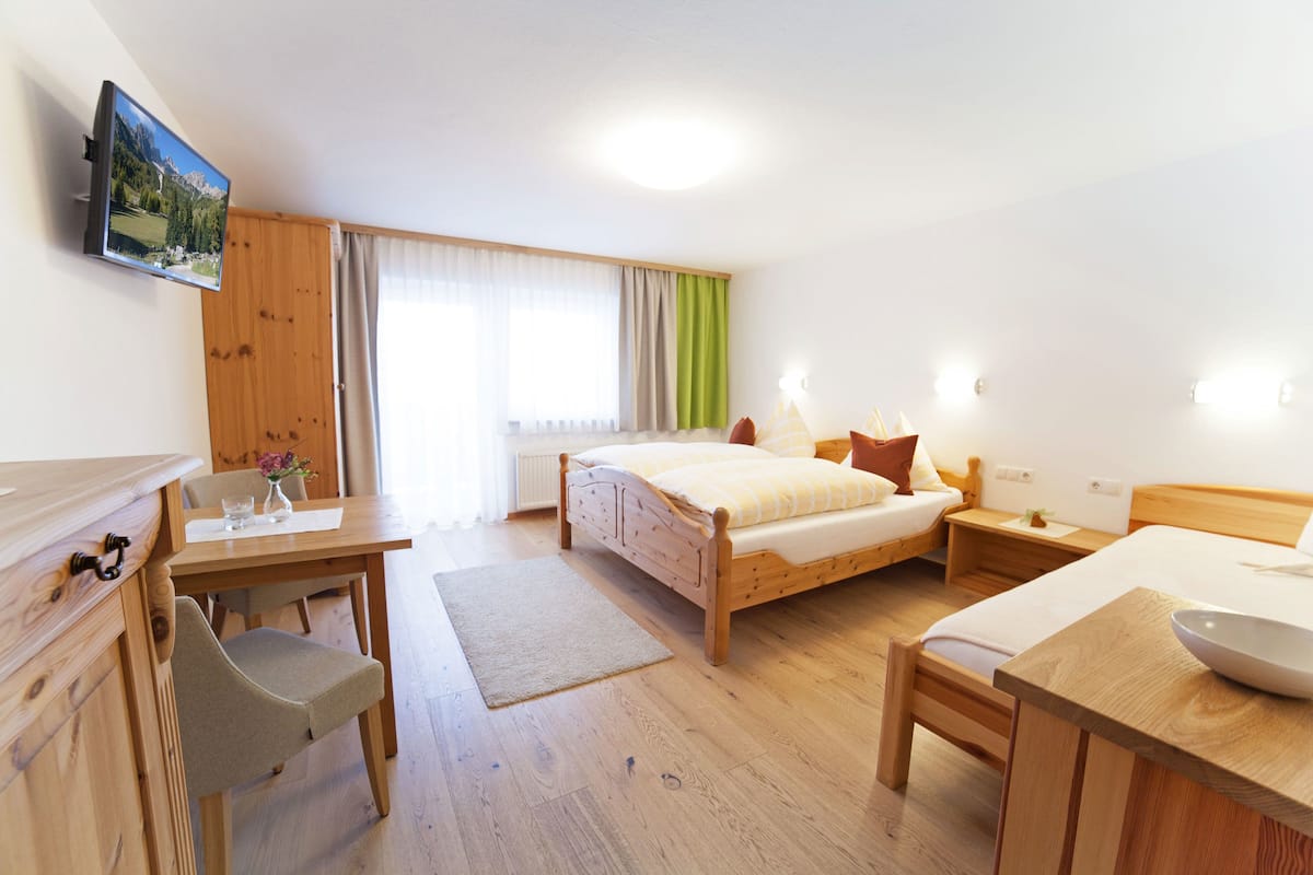 Quarto de hotel com uma cama de madeira, uma escrivaninha com cadeira, uma mesinha com uma tigela e uma televisão na parede.