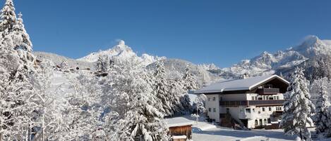 A snow-covered mountain lodge with a large building, surrounded by snow-laden trees and mountains.