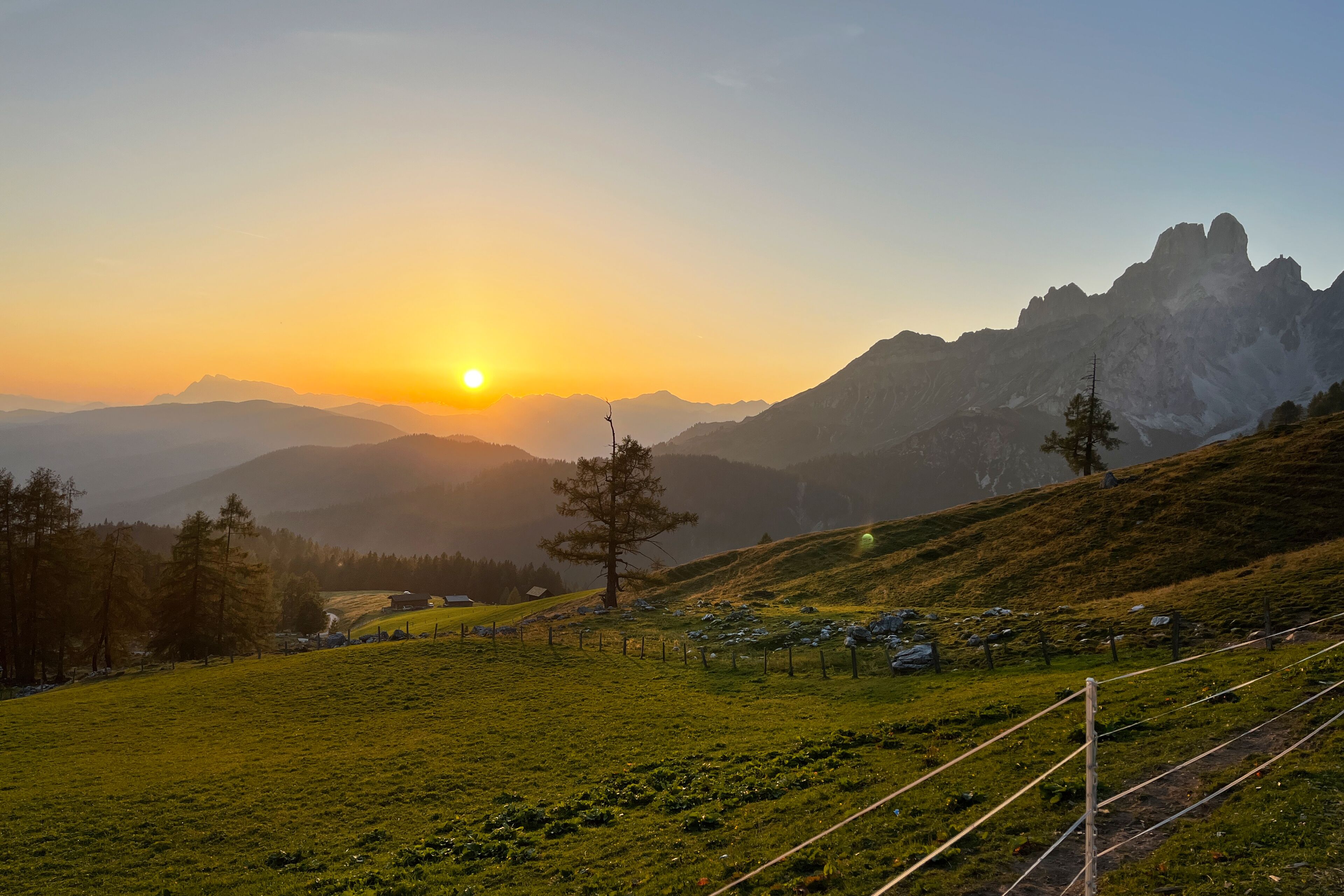 Un paysage montagneux, avec un coucher de soleil, des champs verdoyants et quelques arbres épars.