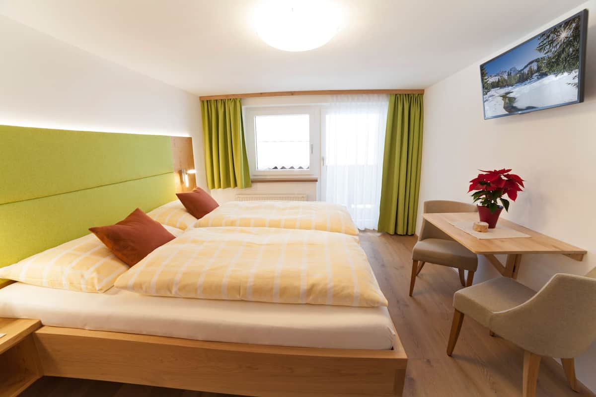 Quarto de hotel com duas camas, cabeceira de madeira, uma mesa com cadeiras, uma planta em vaso e uma janela com cortinas.