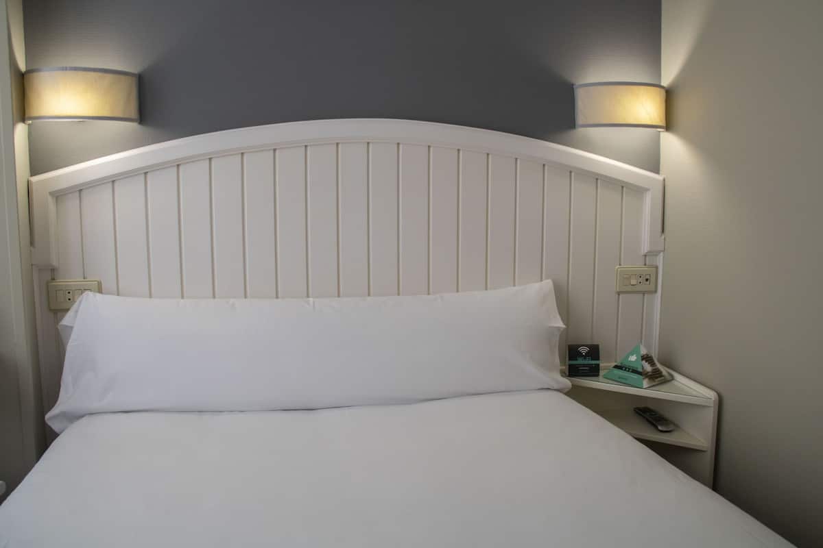 Una cama bien hecha con ropa blanca, un cabecero blanco y dos lámparas fijadas a la pared a cada lado.