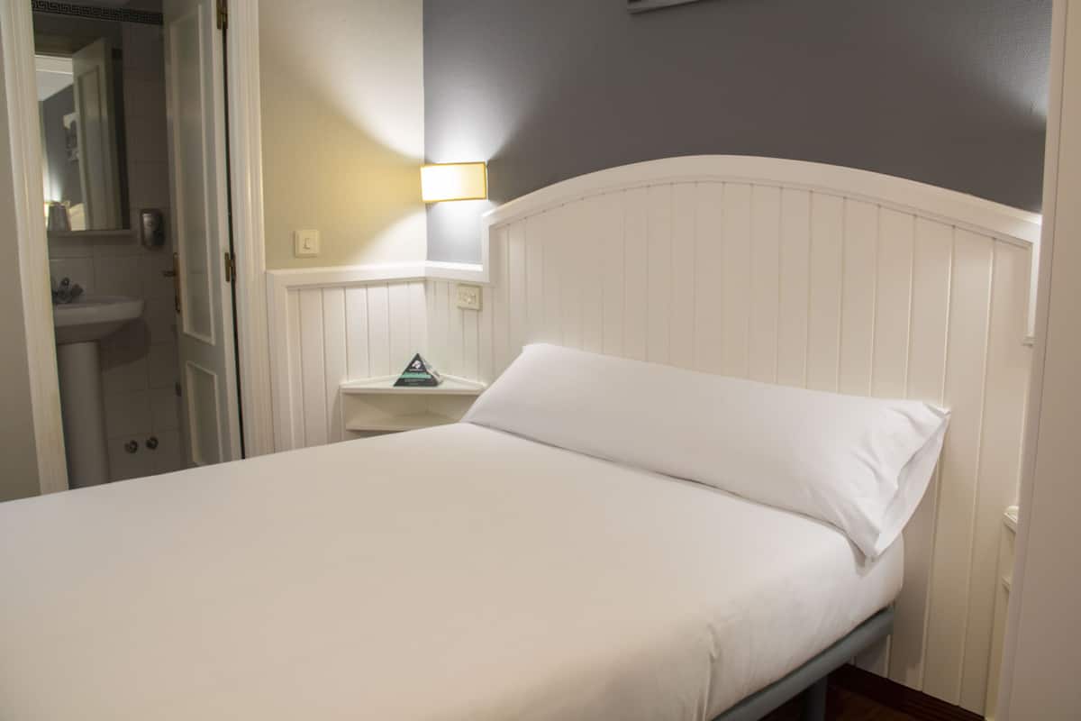 Una cama con ropa de cama blanca, una mesita de noche con lámpara y un baño con espejo y lavamanos.