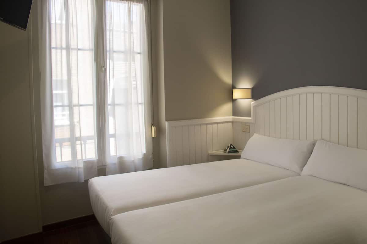 Una habitación de hotel con una cama, una mesita de noche, una lámpara y una ventana con cortinas.