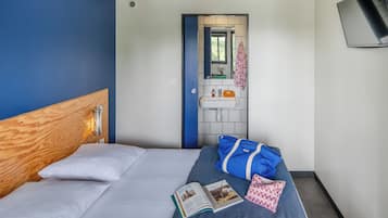 Habitación de hotel con cabecera de madera, cama individual con ropa de cama blanca, una manta azul, un bolso azul, un libro y una mesita de noche pequeña con una lámpara.