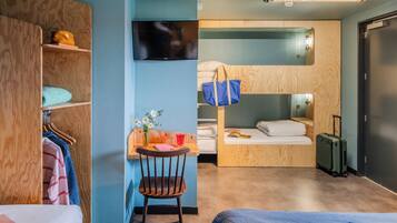 Quarto de hotel compacto com beliche, escrivaninha com cadeira, televisão e mala.