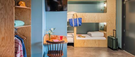 Quarto de hotel compacto com beliche, escrivaninha com cadeira, televisão e mala.