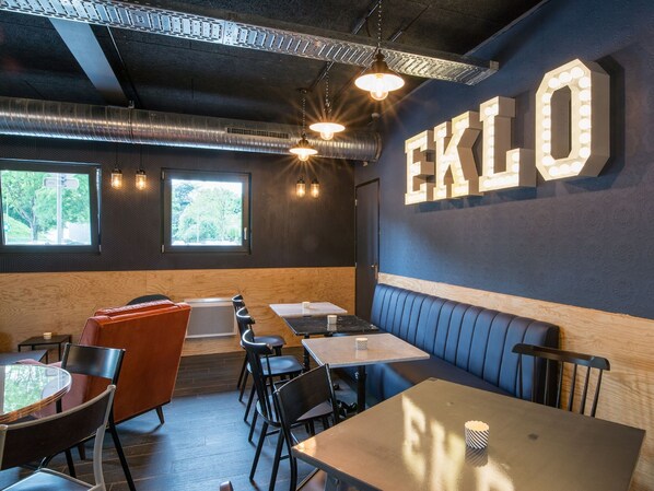 A restaurant interior with a dark wood ceiling, exposed ductwork, and a large illuminated sign reading "EKLO". Tables and chairs are arranged for dining. - Eklo Hotels Lille (Lille)
