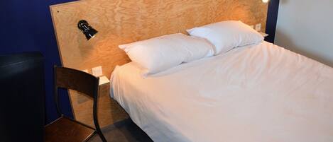 Habitación de hotel con cabecera de madera, cama individual con ropa blanca, una silla y una mesita de noche con lámpara.