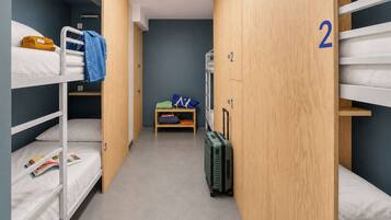 A dormitory room with bunk beds, a small desk, a suitcase, and a wardrobe with numbered doors.