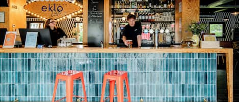 Un bar con mostrador de madera, dos taburetes rojos y un cartel que dice 'Bienvenue eklo bar'.