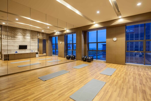 Un gimnasio espacioso con suelo de madera, colchonetas de ejercicio, un televisor montado en la pared y amplias ventanas con vistas a la ciudad.
