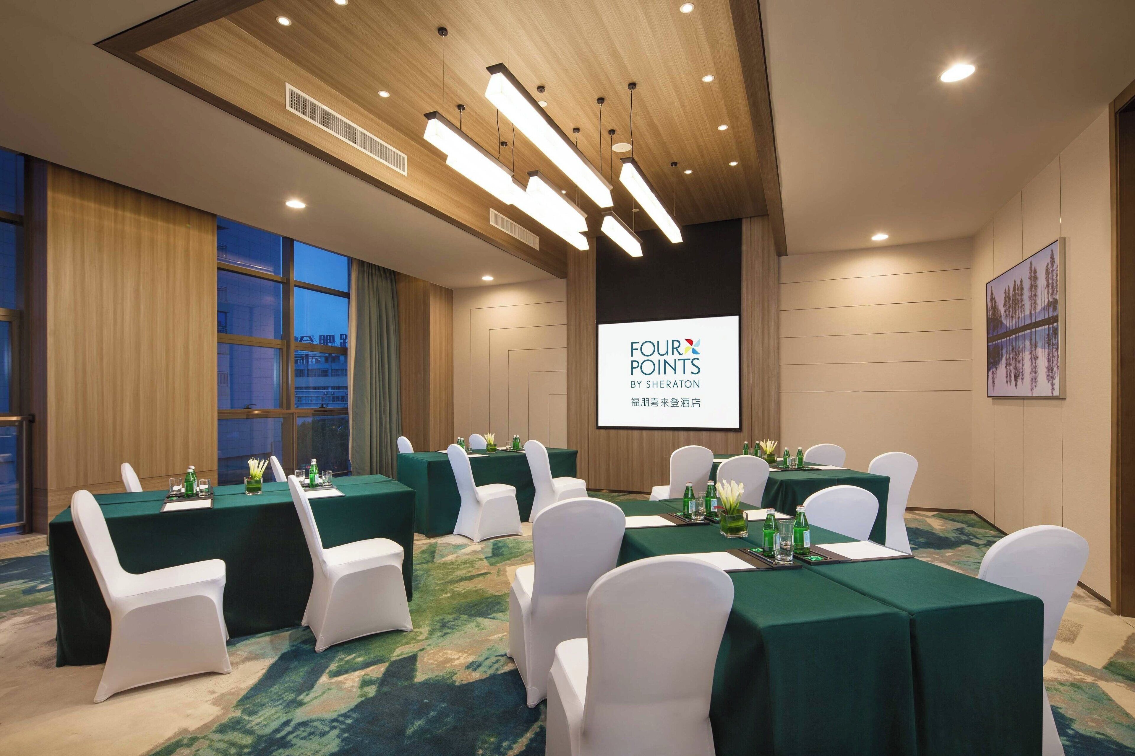 Ett konferensrum med flera bord, vita stolar och en projektorduk som visar texten ”FOUR POINTS BY SHERATON”.