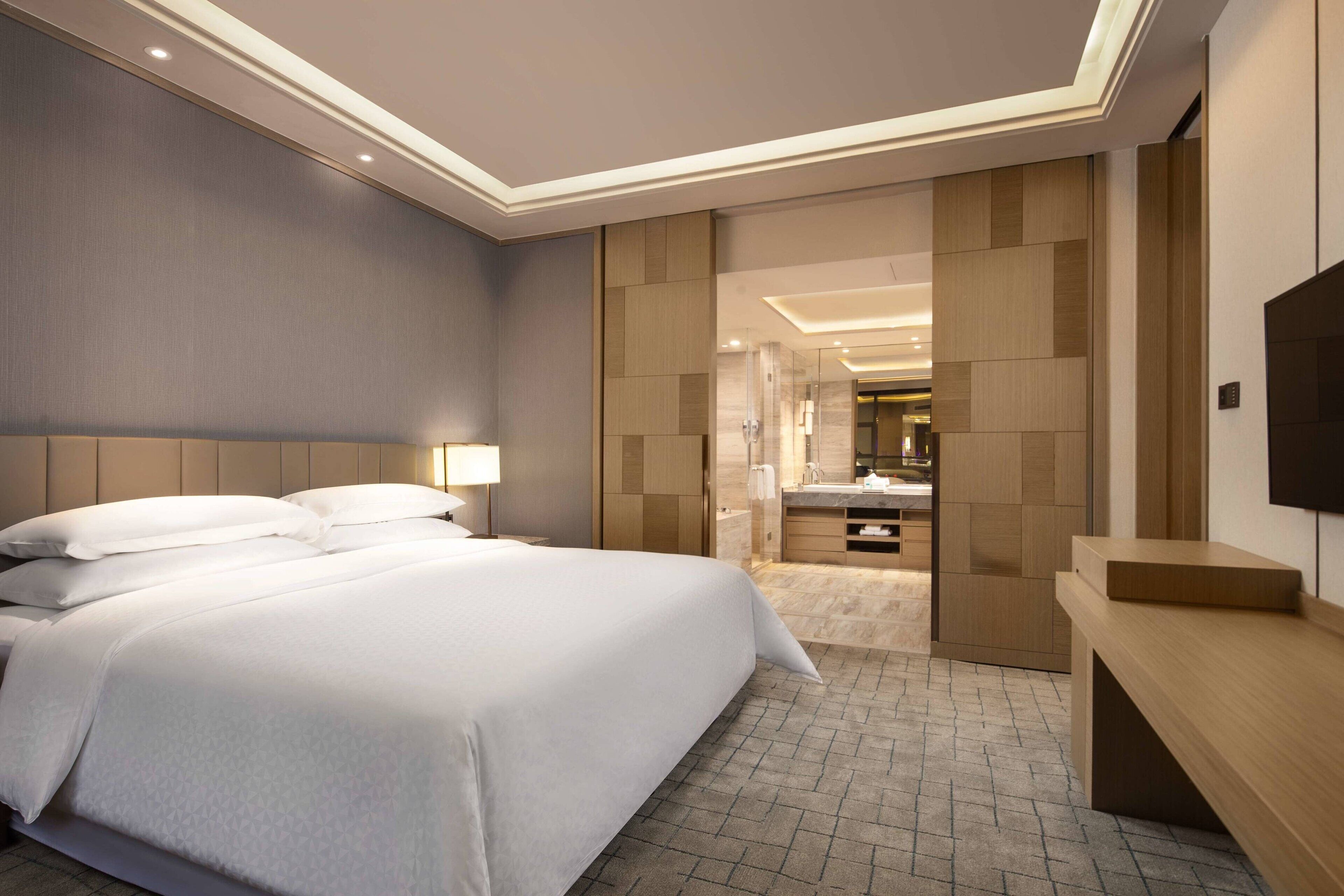 A modern hotel room with a large bed, a bedside table, a TV, and a bathroom visible through an open door.