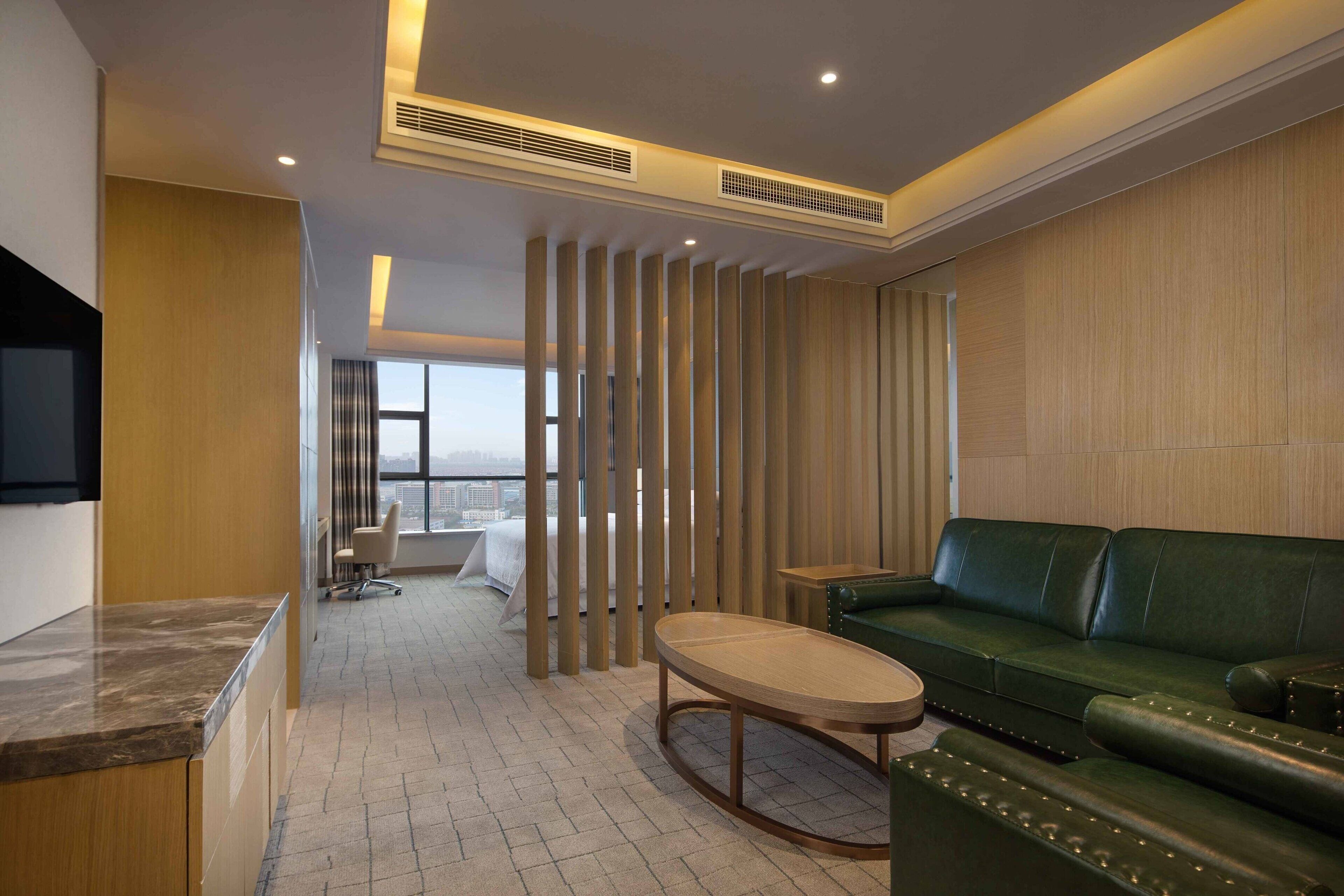 Une chambre d’hôtel moderne avec un canapé en cuir vert, une table basse en bois et une vue sur la ville grâce à de grandes fenêtres.